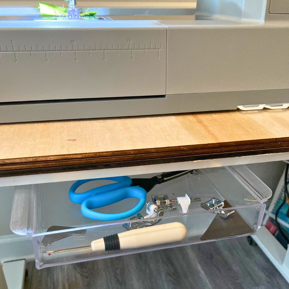 A clear plastic drawer pulled out beneath a sewing machine on a wooden table, holding blue-handled scissors, sewing presser feet and a seam ripper.