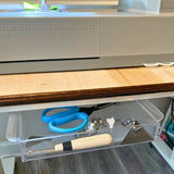 A clear plastic drawer pulled out beneath a sewing machine on a wooden table, holding blue-handled scissors, sewing presser feet and a seam ripper.